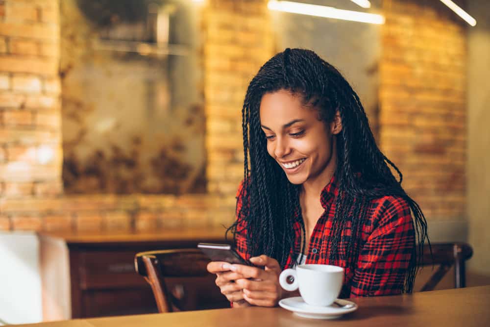 A woman sits in a coffee shop with a drink in front of her. She smiles while on her phone.