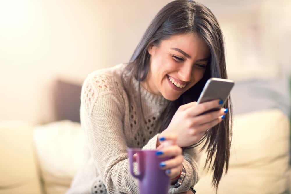 A woman laughs while reading a message on her phone. She\'s holding a purple mug.