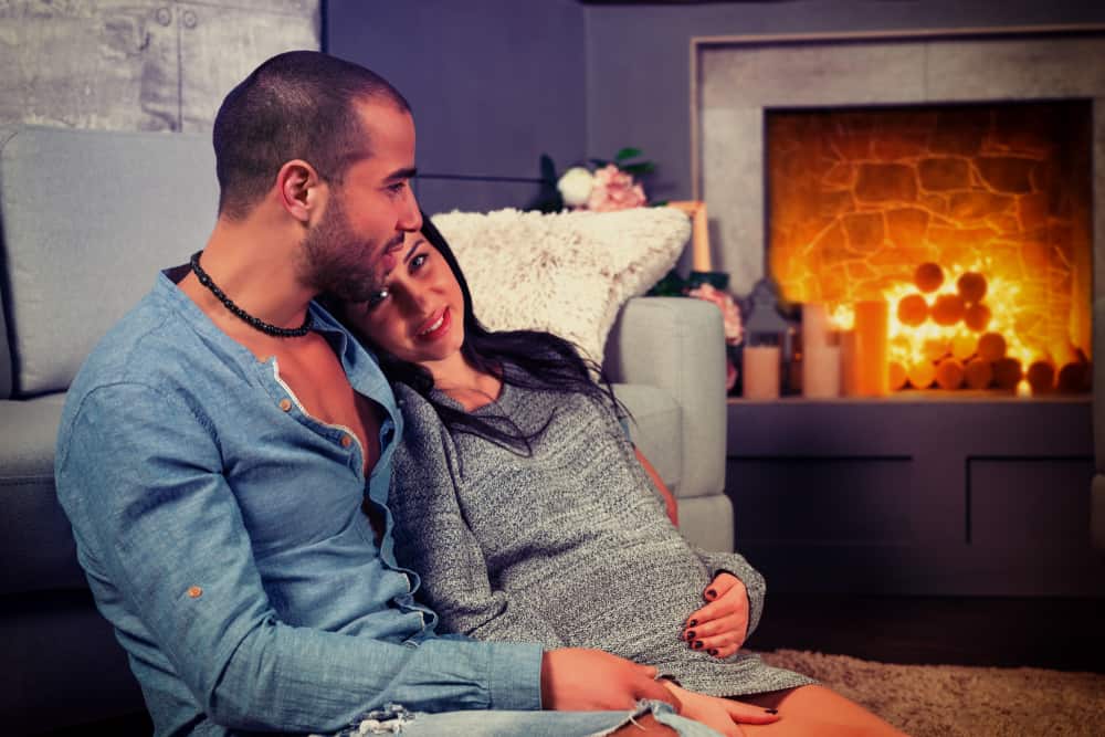 A pregnant woman cuddles by a man indoors next to the fireplace.