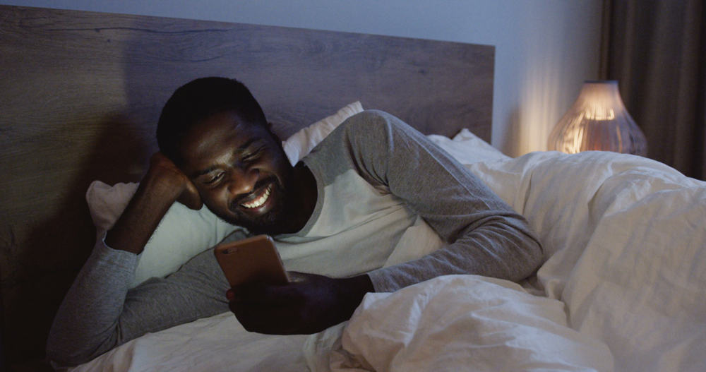 Man laying in bed smiling and reading messages on his phone. 