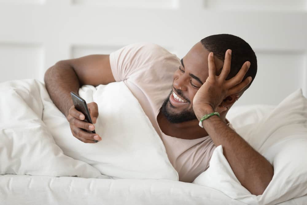 A man laying in bed smiling as he reads something on his phone.