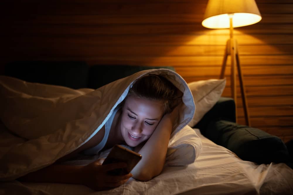 A woman lays in bed with a blanket over her head while reading a message on her phone.