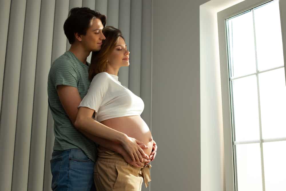 A man holds his pregnant wife\'s belly as they look out the window.