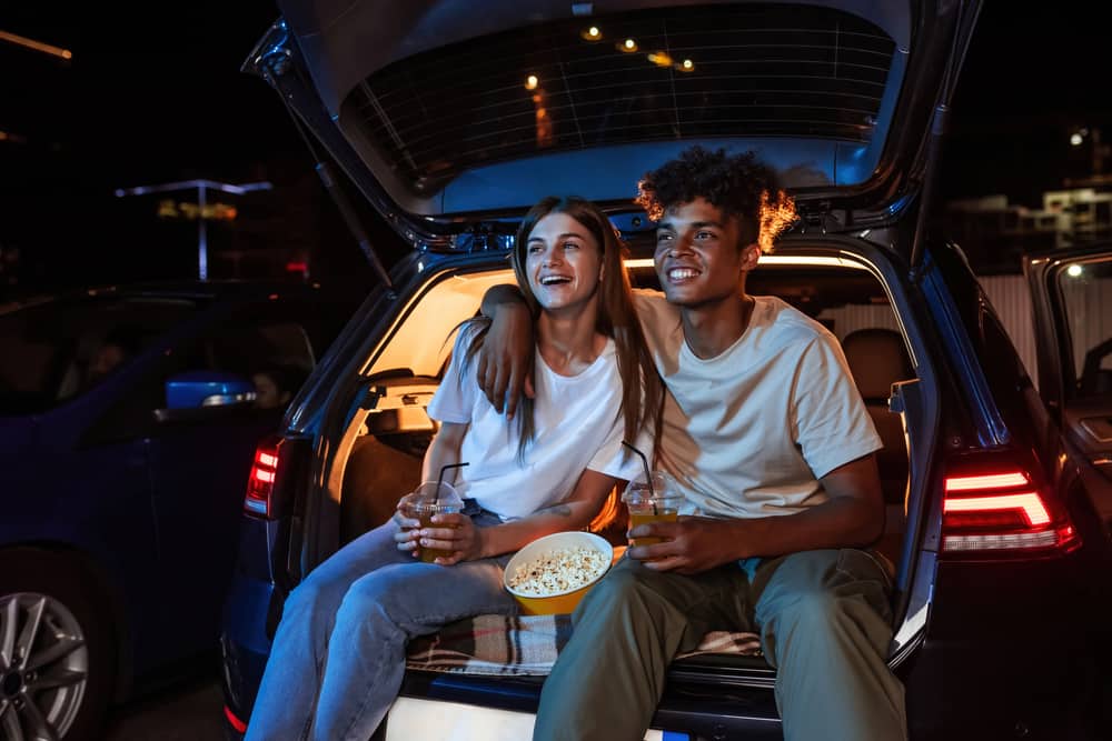 A couple sits in the back of a car holding drinks while popcorn sits between them.