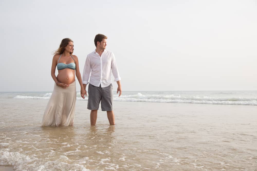 A couple holds hands on the beach. The woman is pregnant.