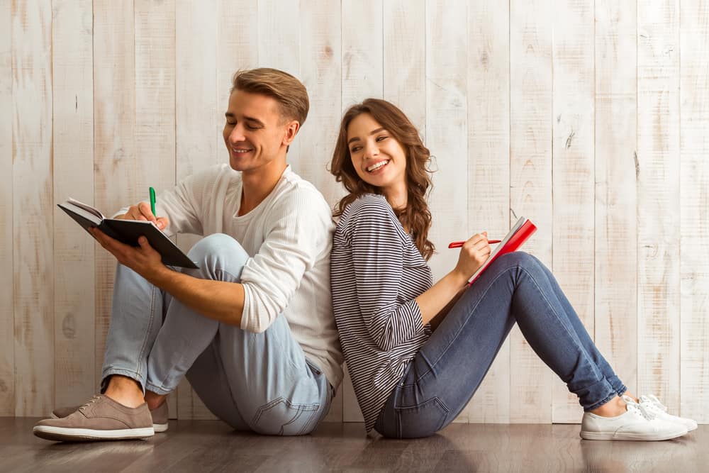 A couple sits back to back while writing in journals, laughing.