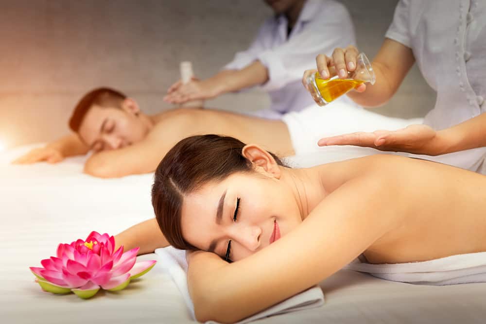 A couple sits inside on their stomach with their eyes closed while receiving a massage.