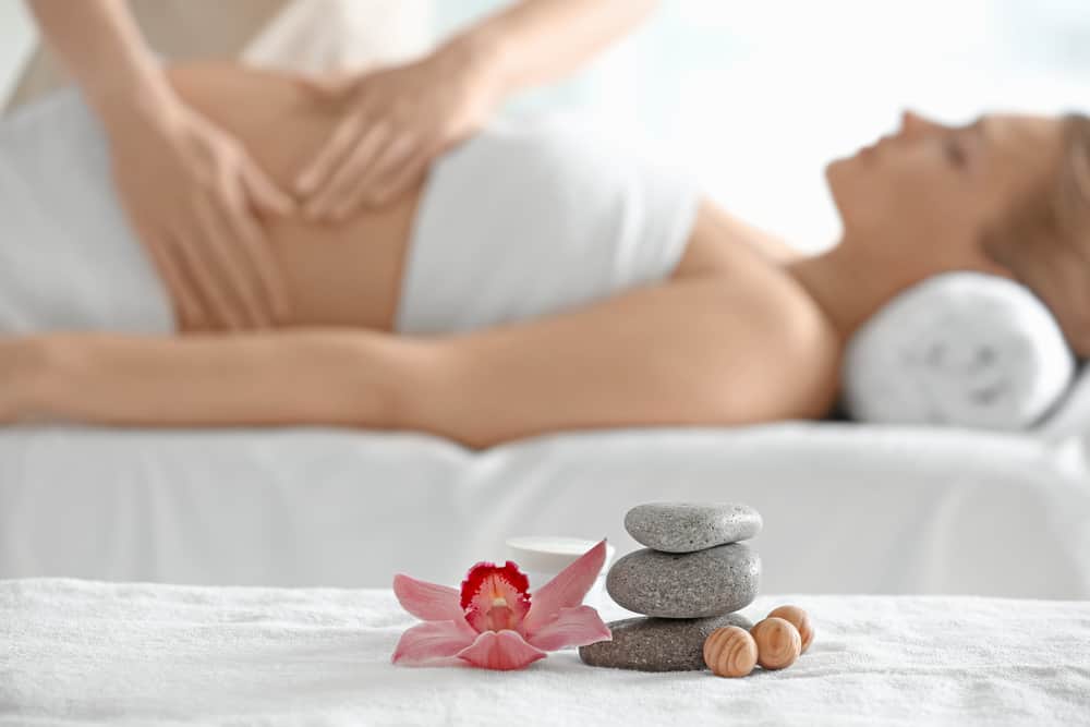 A pregnant woman receives a massage. In the forefront, stones are stacked next to a flower.