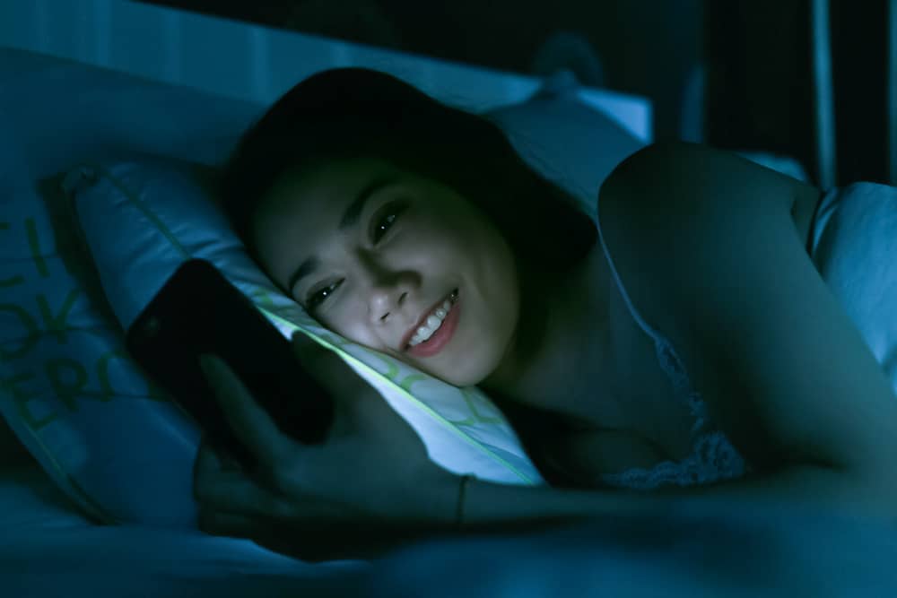 A woman lays in bed smiling while reading a message on her phone.