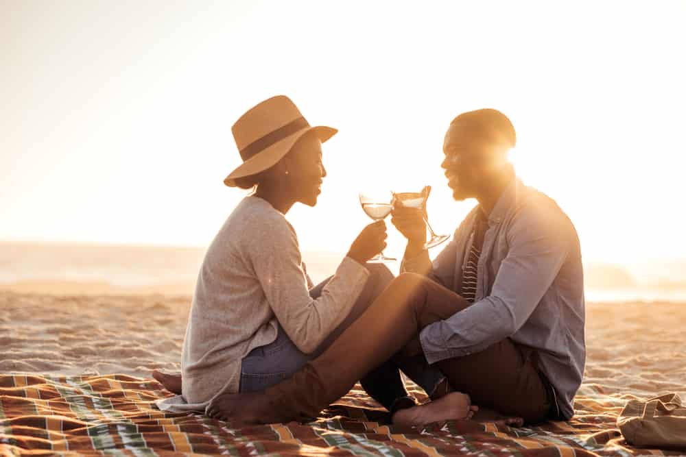 A couple toasts at a beach while the sun sets behind them.
