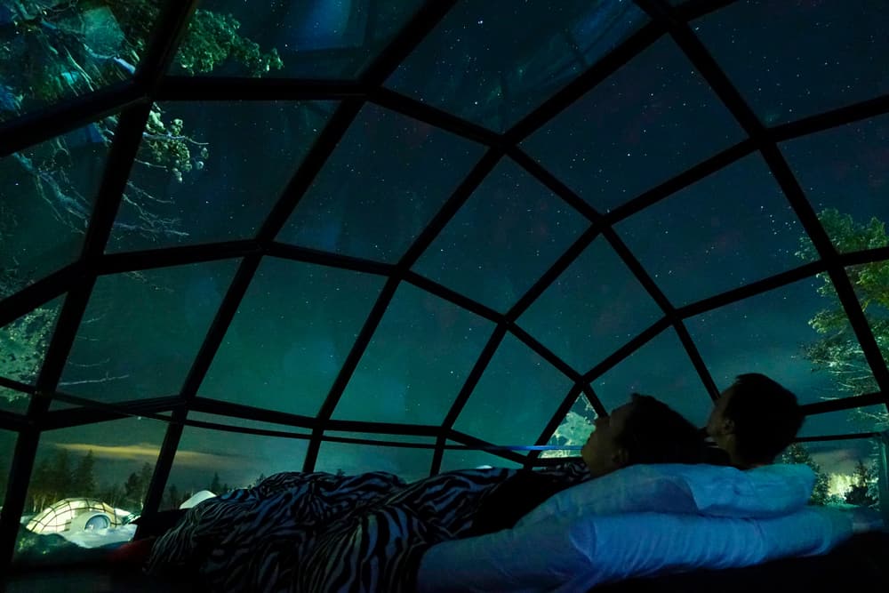 A couple snuggled up together in their glass igloo, looking up at the greenish hued Northern Lights whilom them