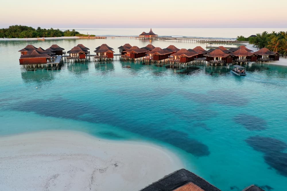 Bright blue water along a white sand beach with a row of bungalows in the water. 