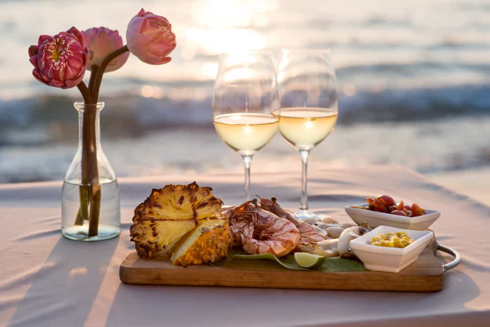 A tray prepared with charcuterie sits next to a glass with flowers and two glasses of wine. The waves crash on the beach behind.