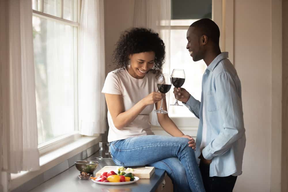 A man and woman laughing in the kitchen drinking wine as sun comes through the windows around them.