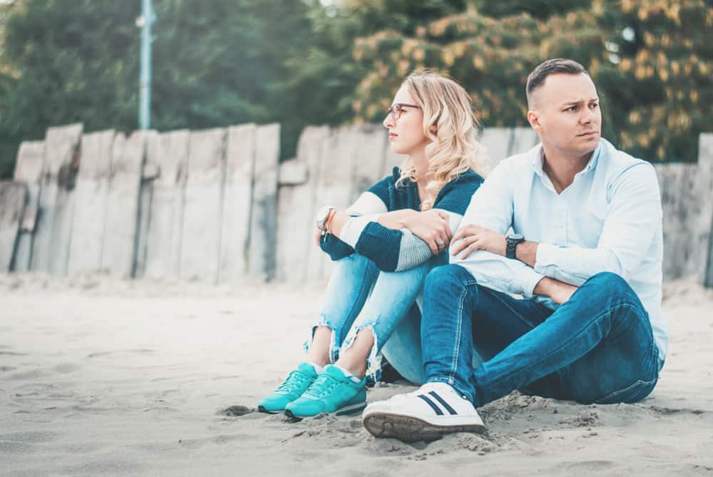 Couple looking unhappy while sitting on the sandy ground in front of a wooden fence.