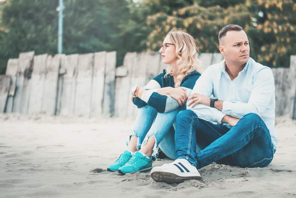 Couple at the looking unhappy in their marriage sitting on the sandy ground in front of a wooden fence