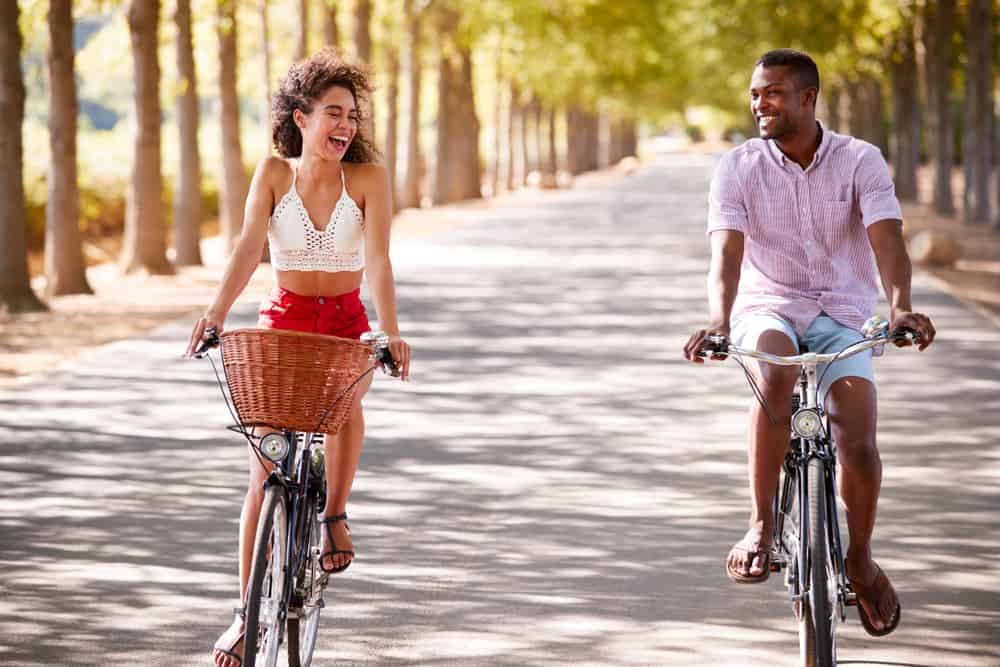 A couple smiling and laughing together as they ride bikes on a wide tree-lined lane with sunlight streaming throug.