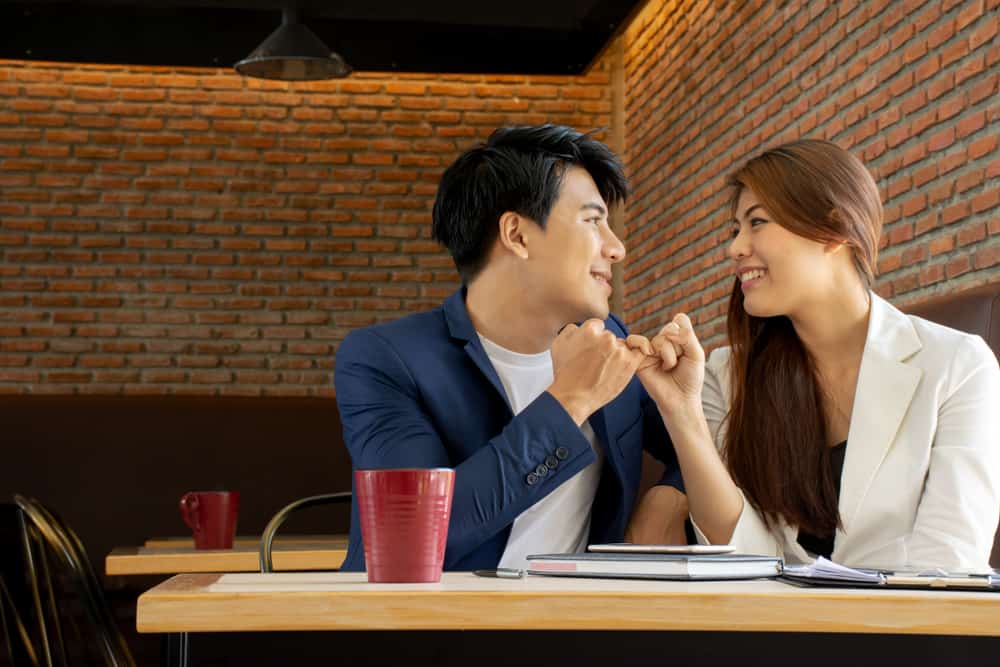 Couple sitting at a table in a restaurant doing a pinky swear while smiling and looking in each other's eyes