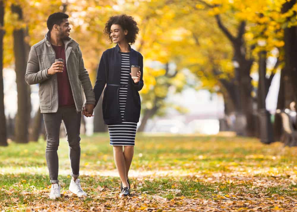 Pareja feliz sonriendo y tomados de la mano mientras pasean por el parque con follaje de otoño detrás de ellos