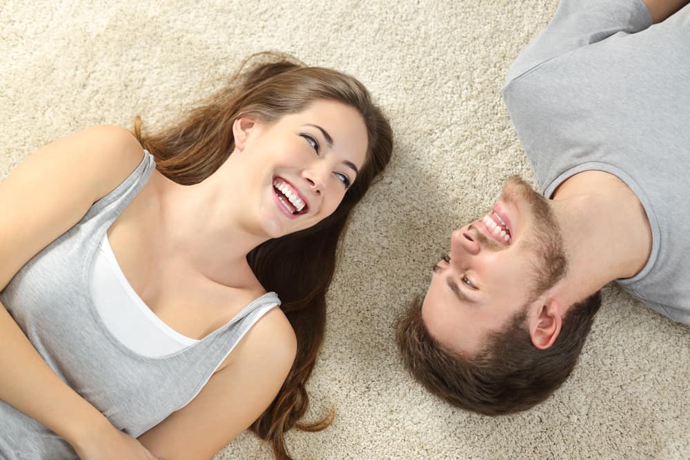 A couple is laying on their back with their heads turned towards each other. They are both smiling.