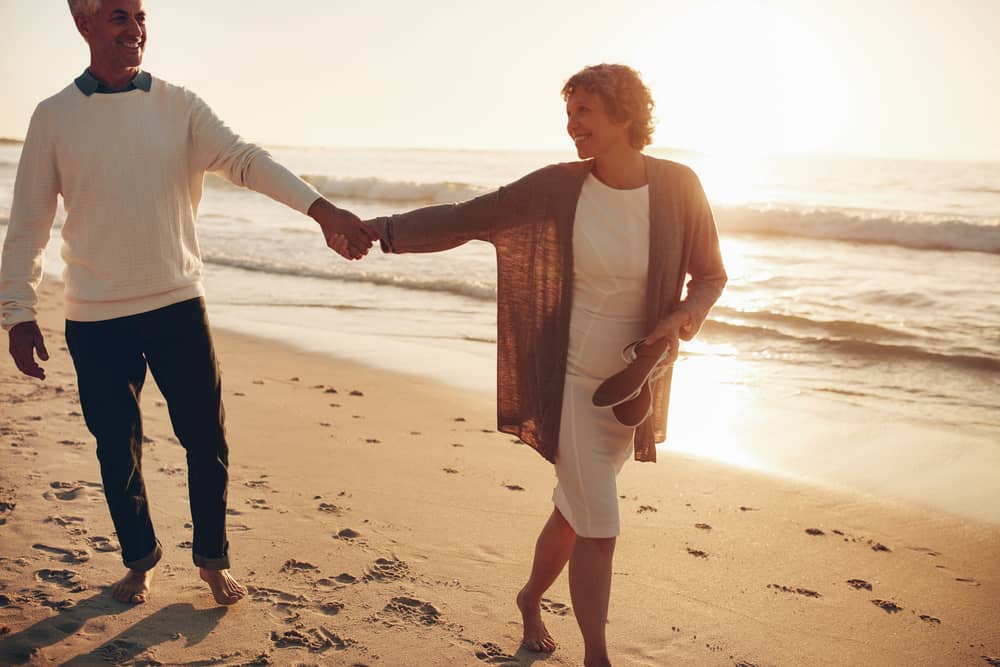 Mature couple holding hands and walking on a beach barefoot at sunset, smiling together with the sea behind them