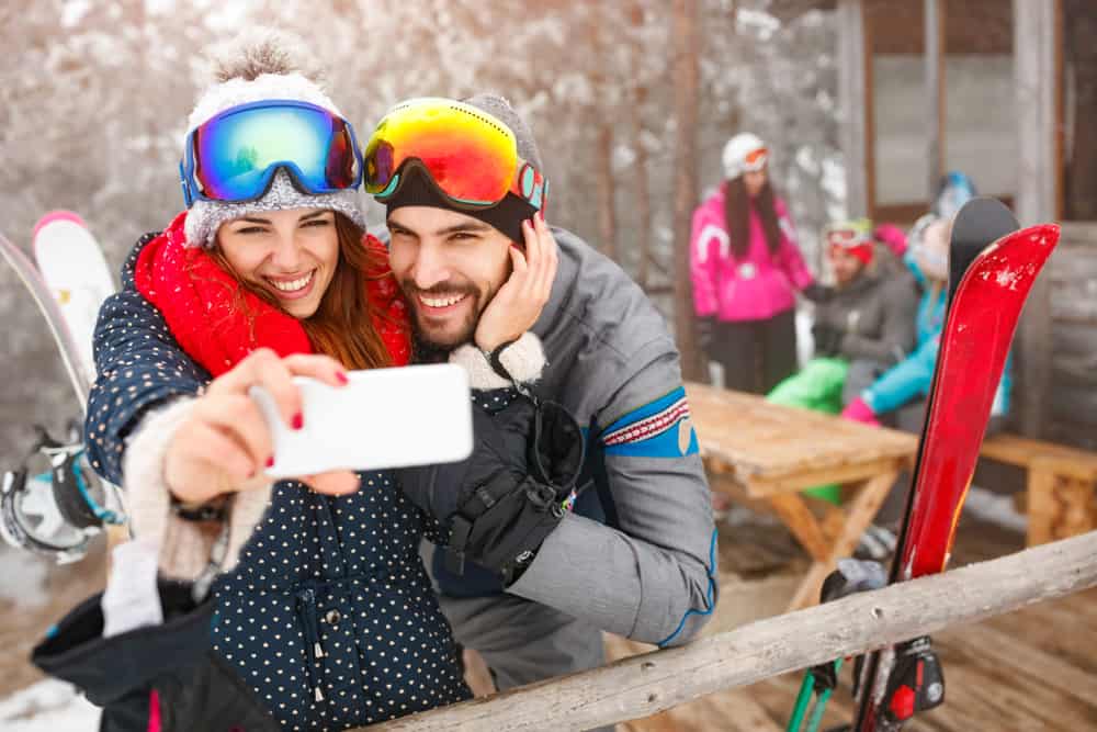 Couple smiling as they take a selfie together wearing ski goggles on their heads at a ski resort