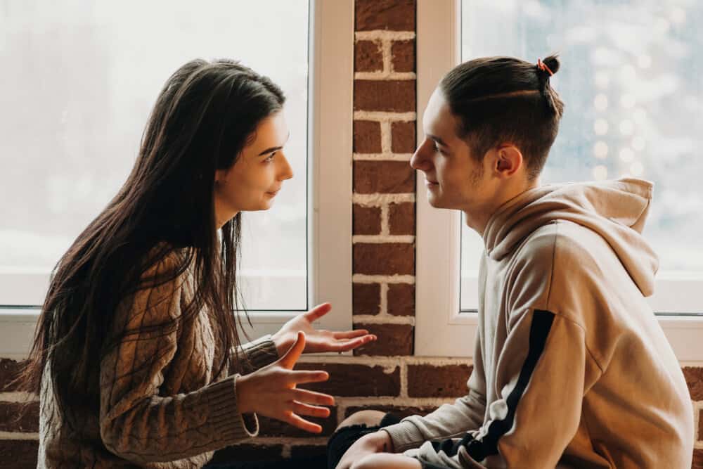 A young couple sits face to face and has an earnest conversation in front of a red brick wall with windows. The woman is moving her hands.