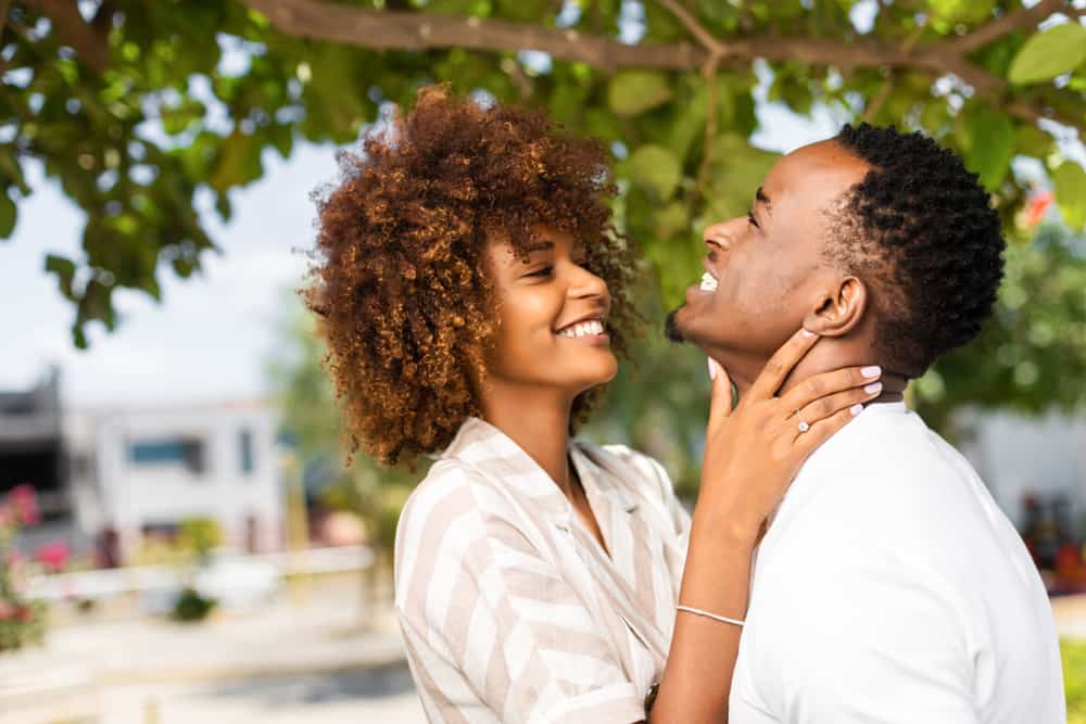 A married couple embraces under a tree as the wife smiles directly at her husband who is happily looking up and grinning