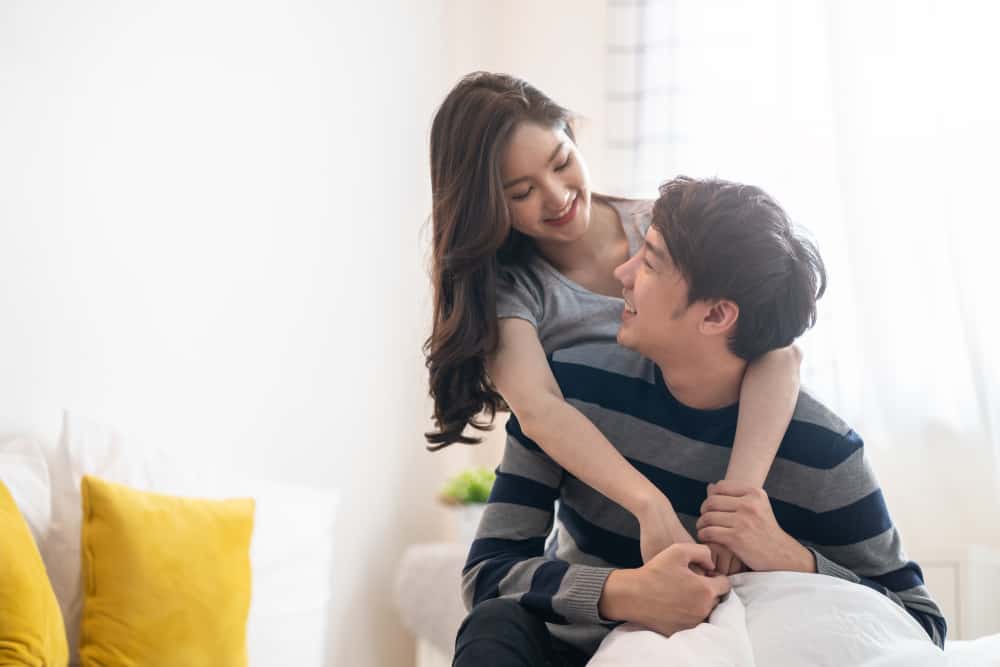 A wife hugs her husband from behind as they both smile at each other in a bright, white room