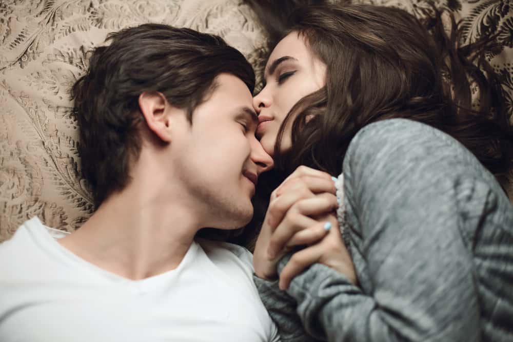 A wife kisses her husband on the forehead as they lay snuggled up together in bed holding hands