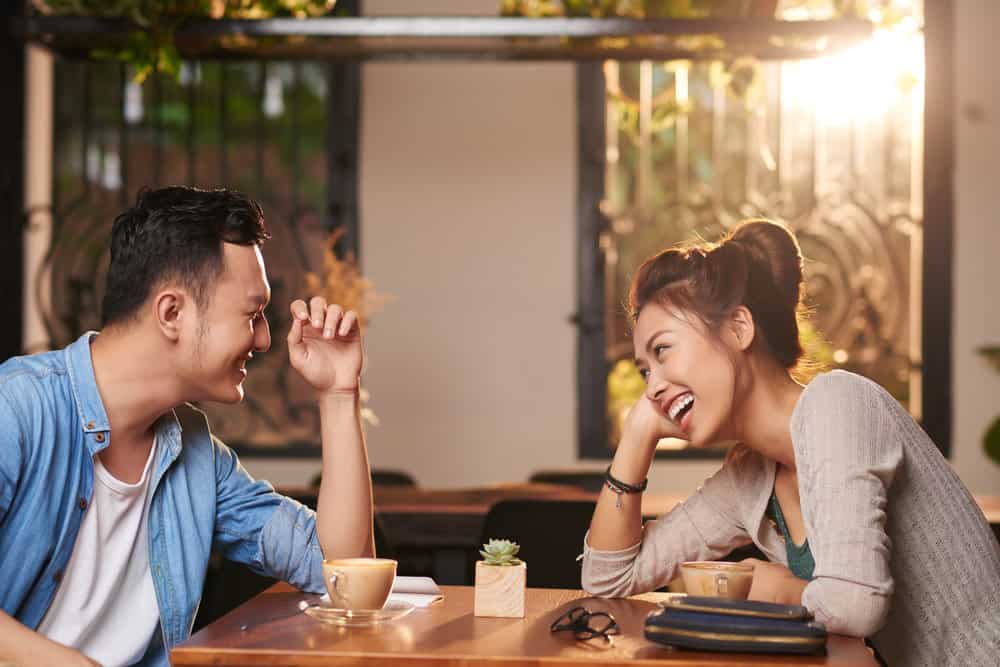 A young couple sits across from each other in a cafe and laughs together during a date. Sunlight pours in through windows behind them.