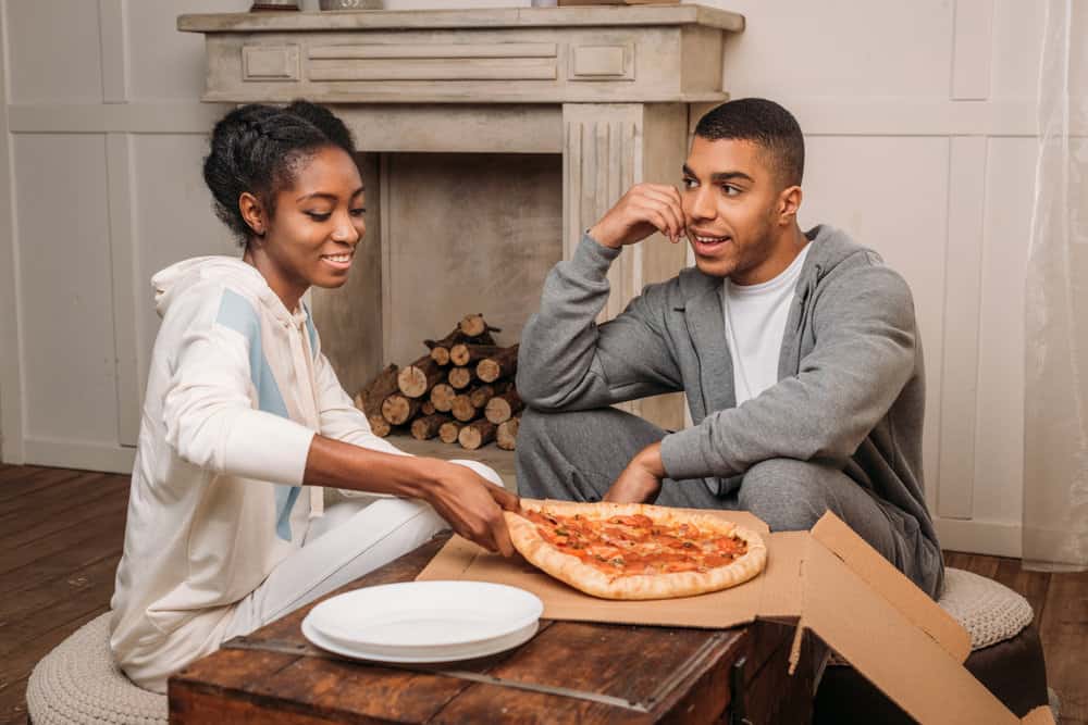 A couple sits on the floor of their home with a pizza between them while smiling.
