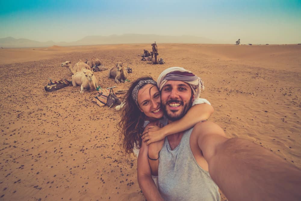 A couple grins as they take a selfie in front of camels on a safari caravan in the desert; a pale blue sky is overhead