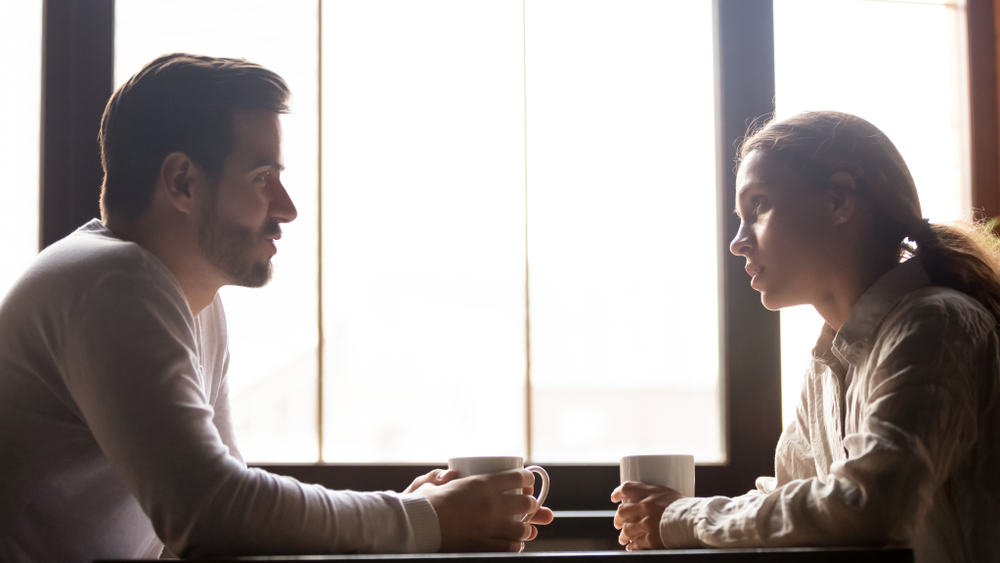 A couple sits across a table with coffee mugs in their hands, having a serious converation