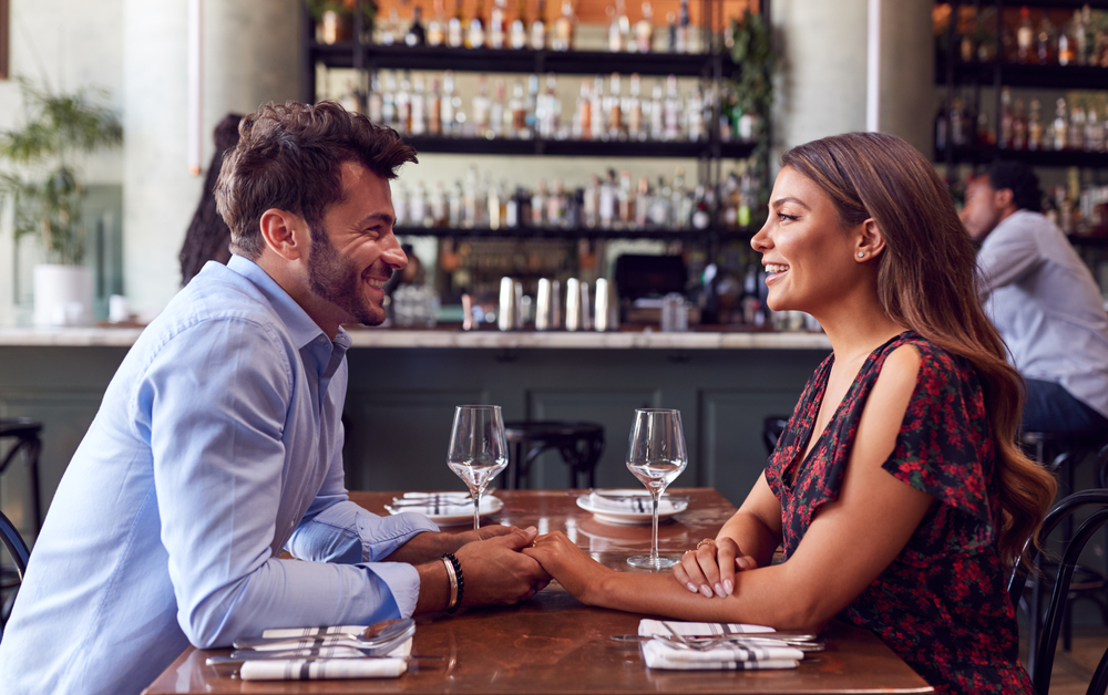 A man and woman sit across from each other in a restaurant, smiling and holding hands.