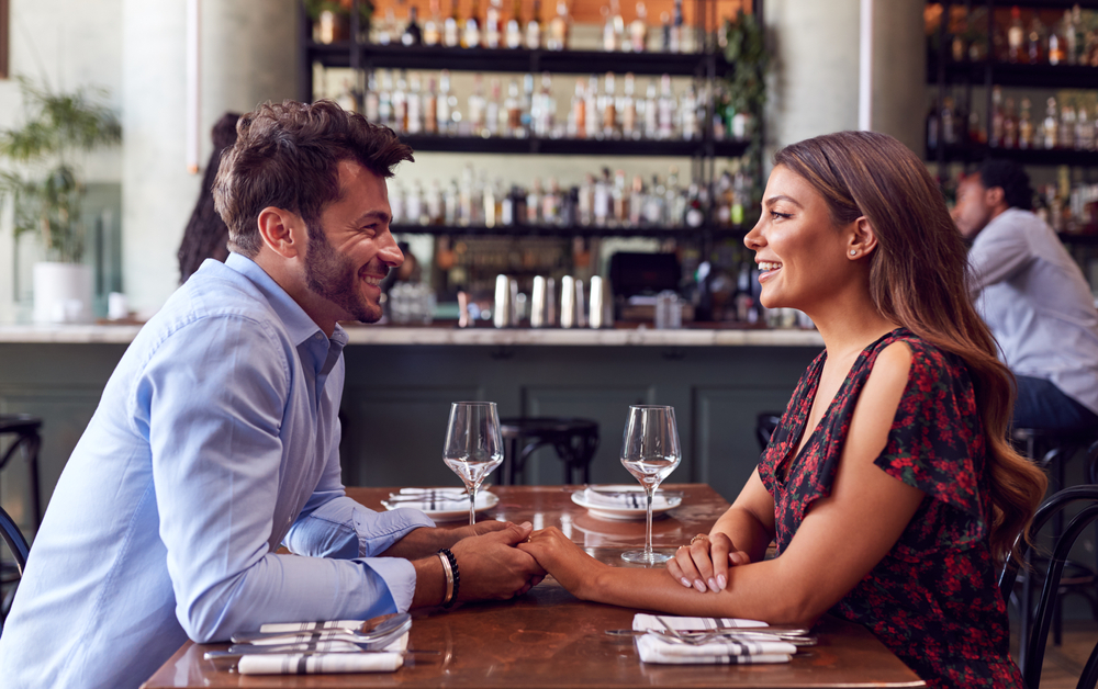 A man and a woman in a new relationship sitting across from each other in a restaurant, smiling and holding hands.