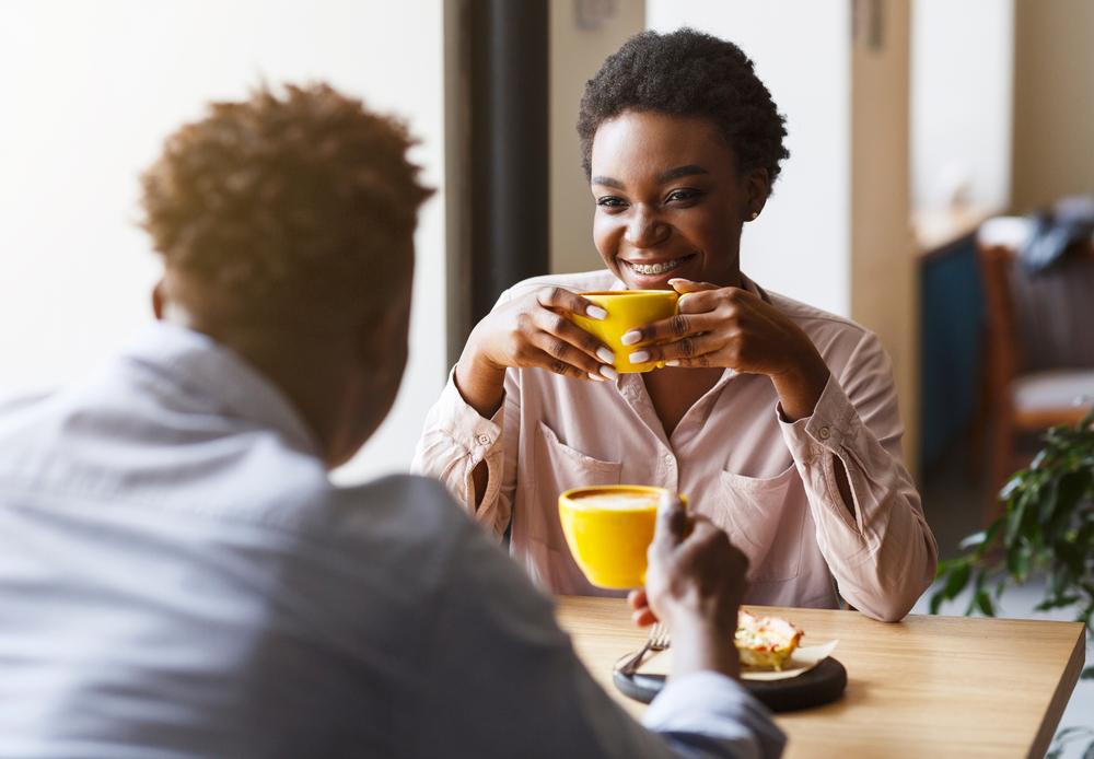 A couple is sitting in across a table from each other, sharing a meal and smiling as sunlight streams through the window beside them