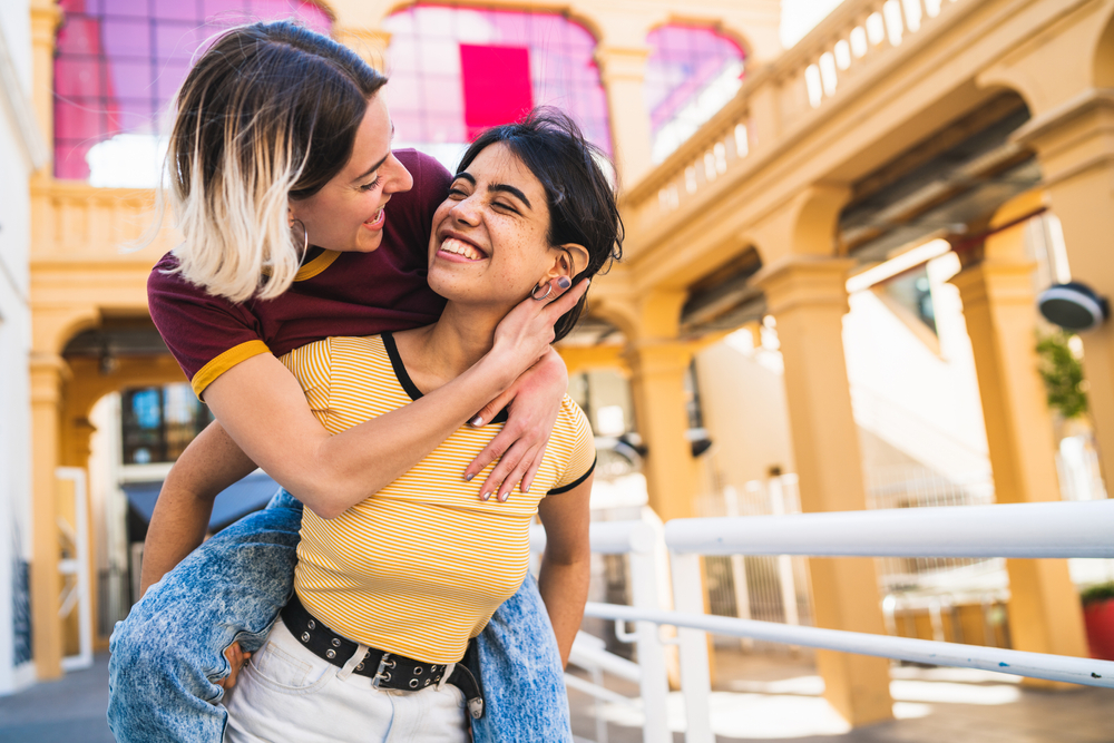 A woman gives her partner a piggyback ride as they smile widely at each other.