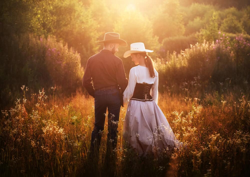 A couple walks hand in hand in cowboy clothing in a forest.