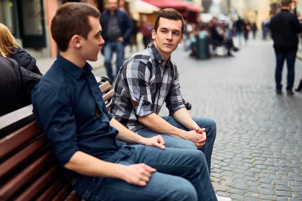 A couple sit on a bench while chatting with one another on a busy street.