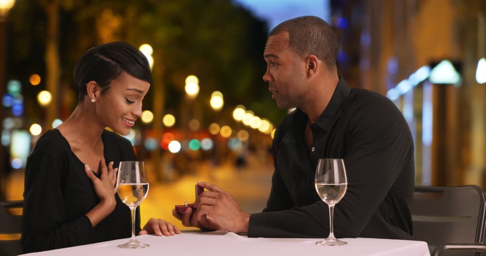 A man proposes to a woman at night. They each have a glass of wine on the table.