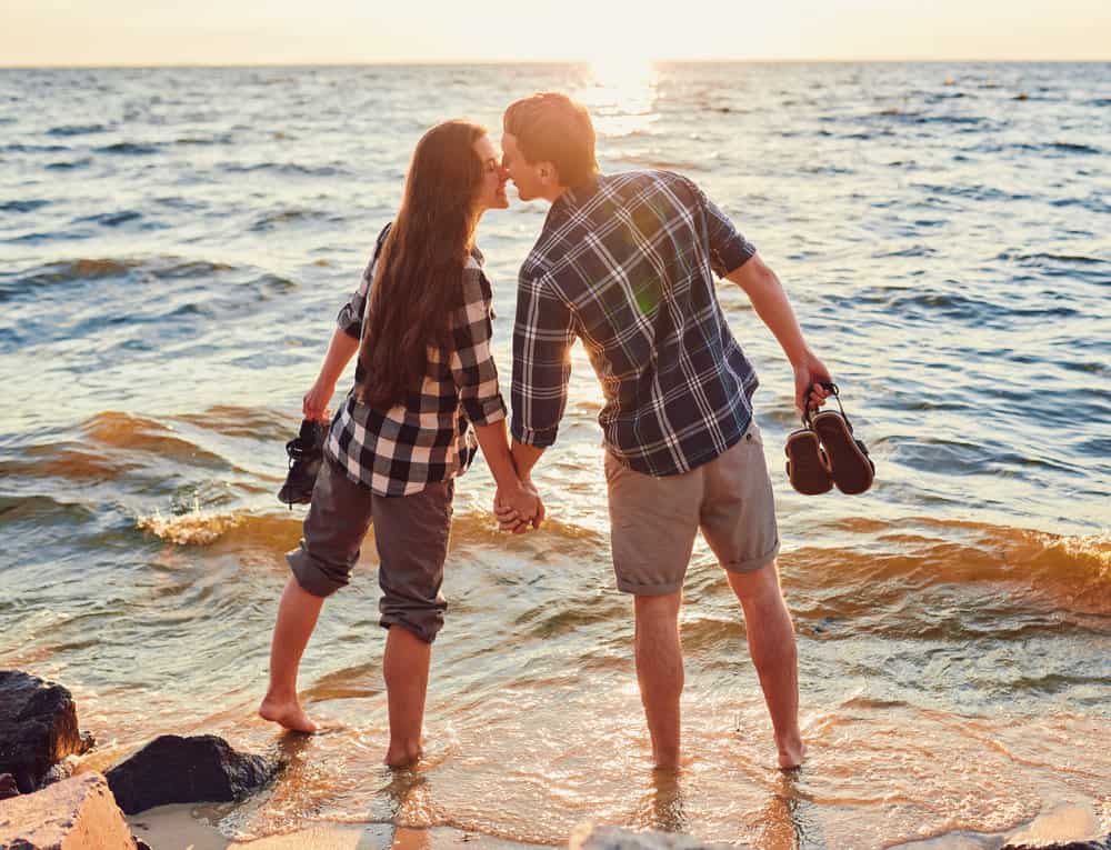 A couple in plaid shirts stands in the water with no shoes on, kissing.