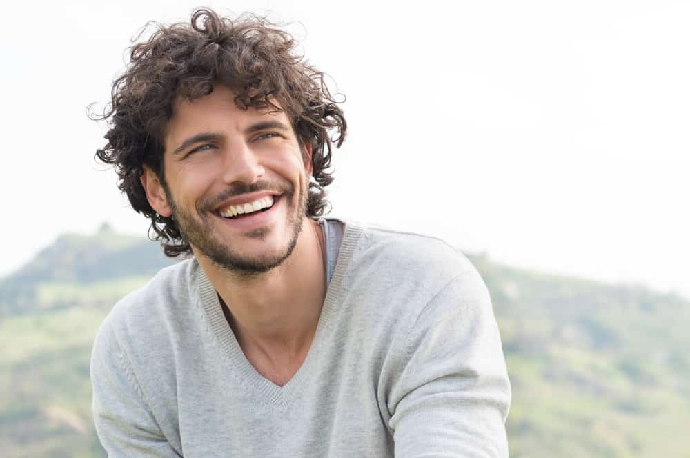 A man smiles outdoors with brown curly hair.