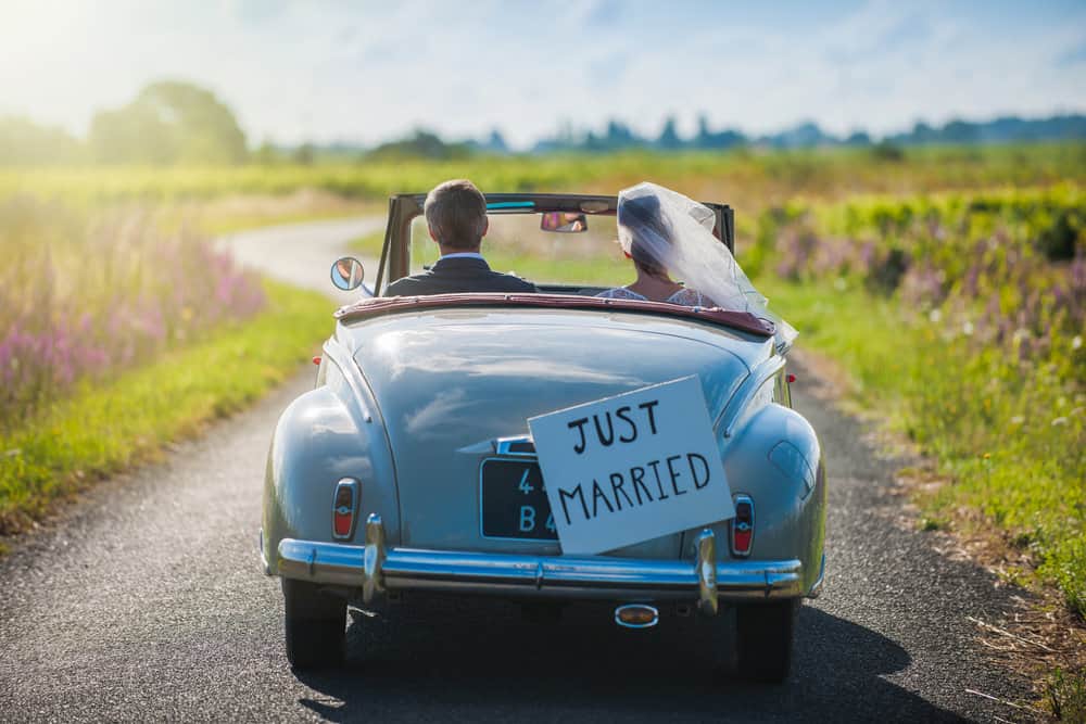 A couple in the first year of marriage drives off in a car with a Just Married sign on the back