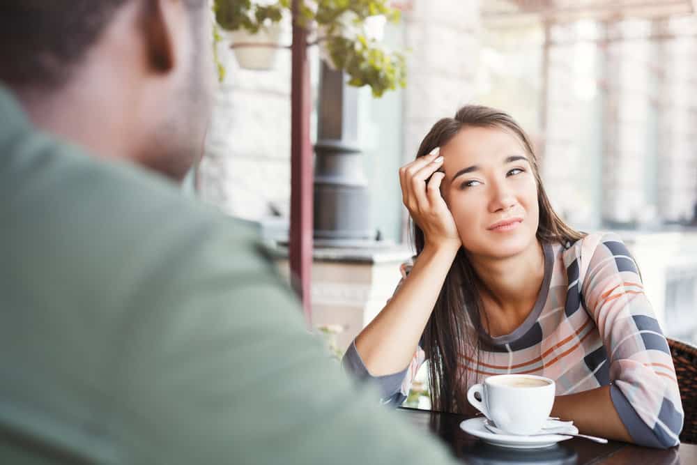 A woman sits at a table with a white mug of coffee in front of her, confused.