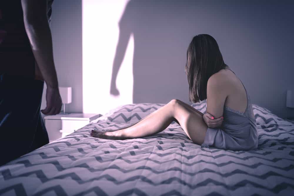 A woman sits sad on her bed, while the silhouette of a man is seen on the wall.