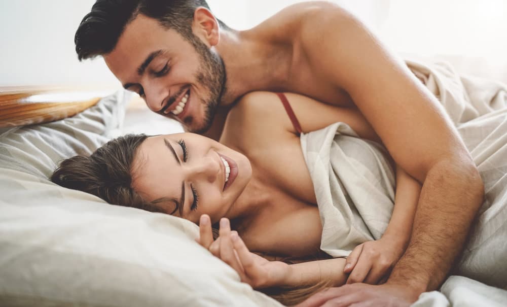 A man cuddles a woman in bed while they both smile.