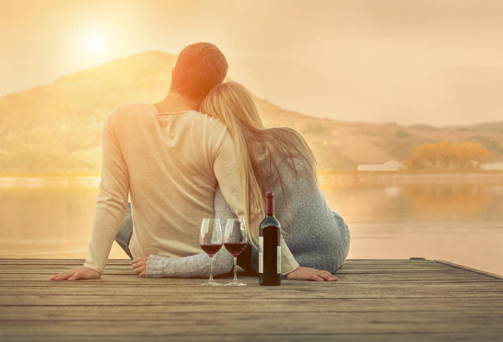 A couple sits together, cuddling into one another, on a lake. Two glasses of red wine and a bottle sit behind them on the pier.