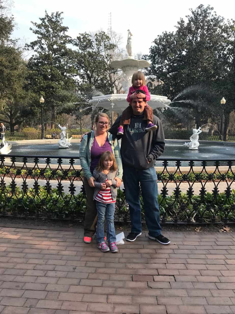 A family of four stands in front of a fountain outdoors.