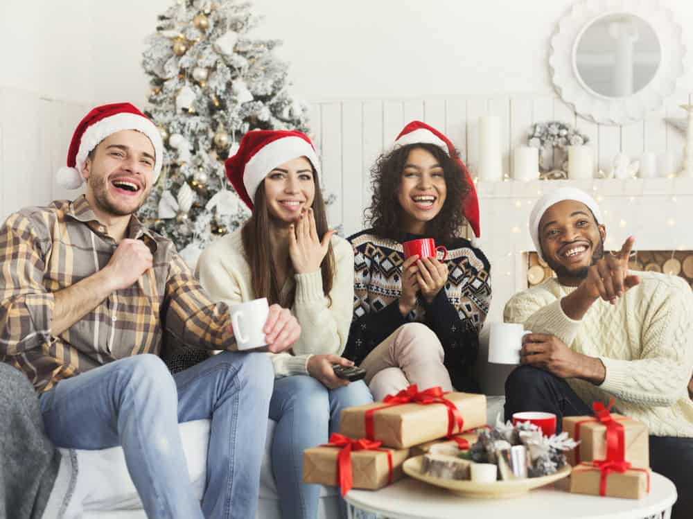 A group of friends with Santa hats on all laugh while watching television. Presents are on the table in front of them. A christmas tree is decorated behind them.